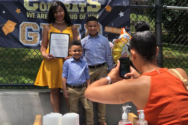 An 8th grader in a yellow dress holds her diploma while her brothers, in blue shirts and slacks, pose with her as their mother takes their picture